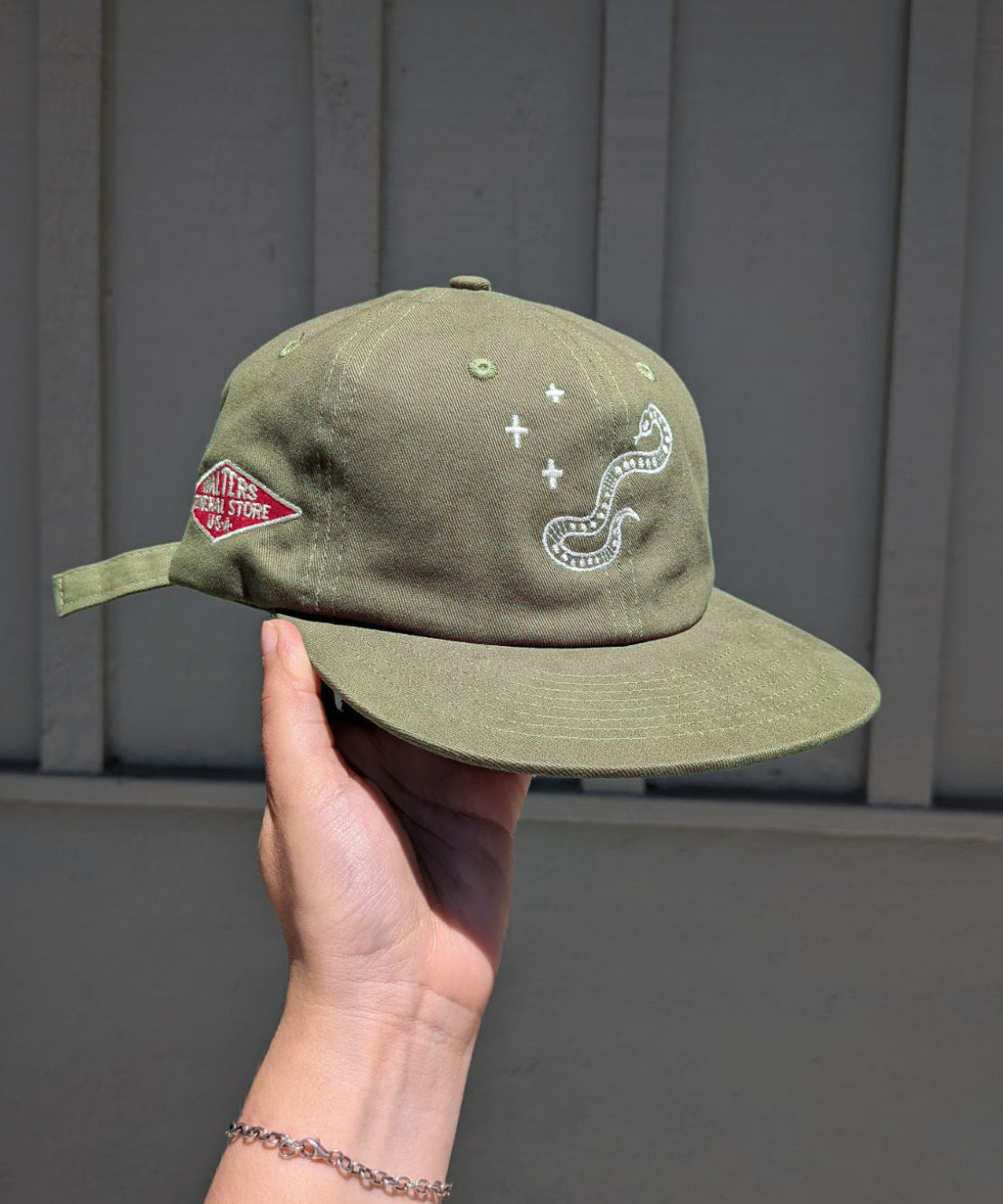 An olive green cap held by a hand against a gray paneled wall. The cap features white embroidered graphics, including a snake and plus sign, and a red diamond Walters General Store logo on the side.