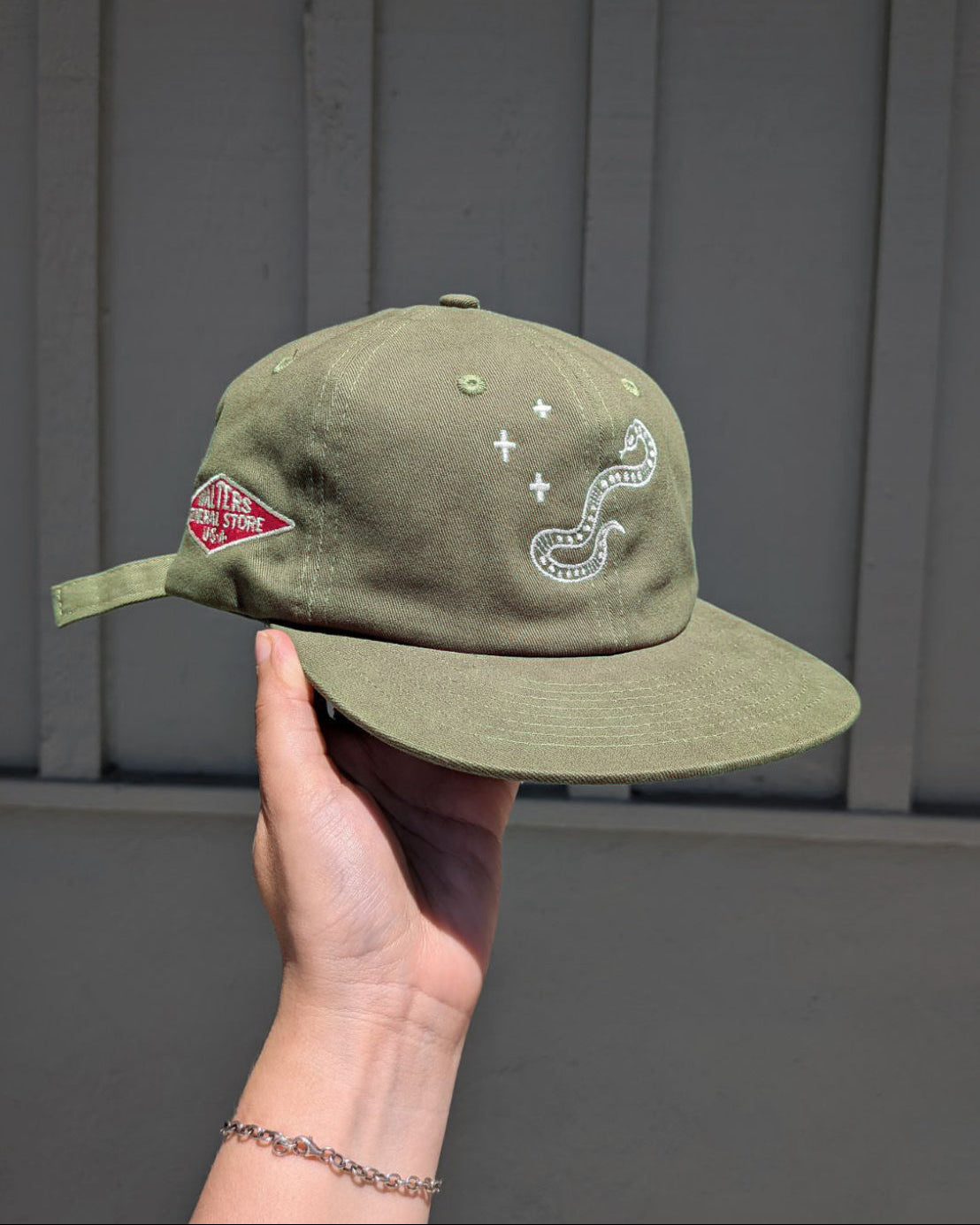 An olive green cap held by a hand against a gray paneled wall. The cap features white embroidered graphics, including a snake and plus sign, and a red diamond Walters General Store logo on the side.