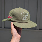 An olive green cap held by a hand against a gray paneled wall. The cap features white embroidered graphics, including a snake and plus sign, and a red diamond Walters General Store logo on the side.