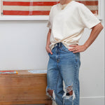 A woman stands indoors wearing a cream short-sleeve t-shirt tucked in at the waist and distressed blue jeans, with an American flag framed on the wall behind her and a wooden chest beside her.