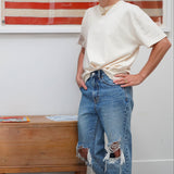A woman stands indoors wearing a cream short-sleeve t-shirt tucked in at the waist and distressed blue jeans, with an American flag framed on the wall behind her and a wooden chest beside her.