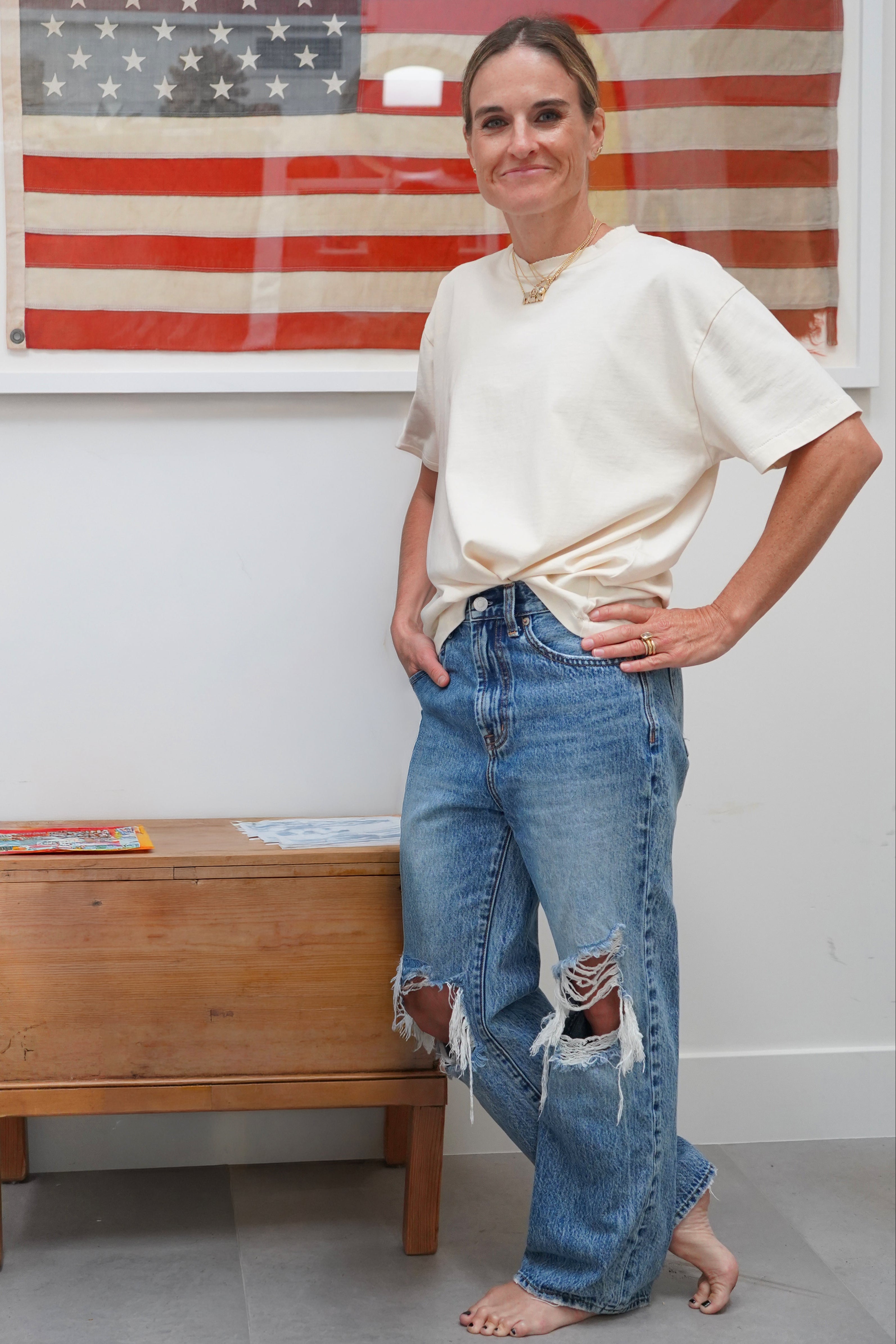 A woman stands indoors wearing a cream short-sleeve t-shirt tucked in at the waist and distressed blue jeans, with an American flag framed on the wall behind her and a wooden chest beside her.