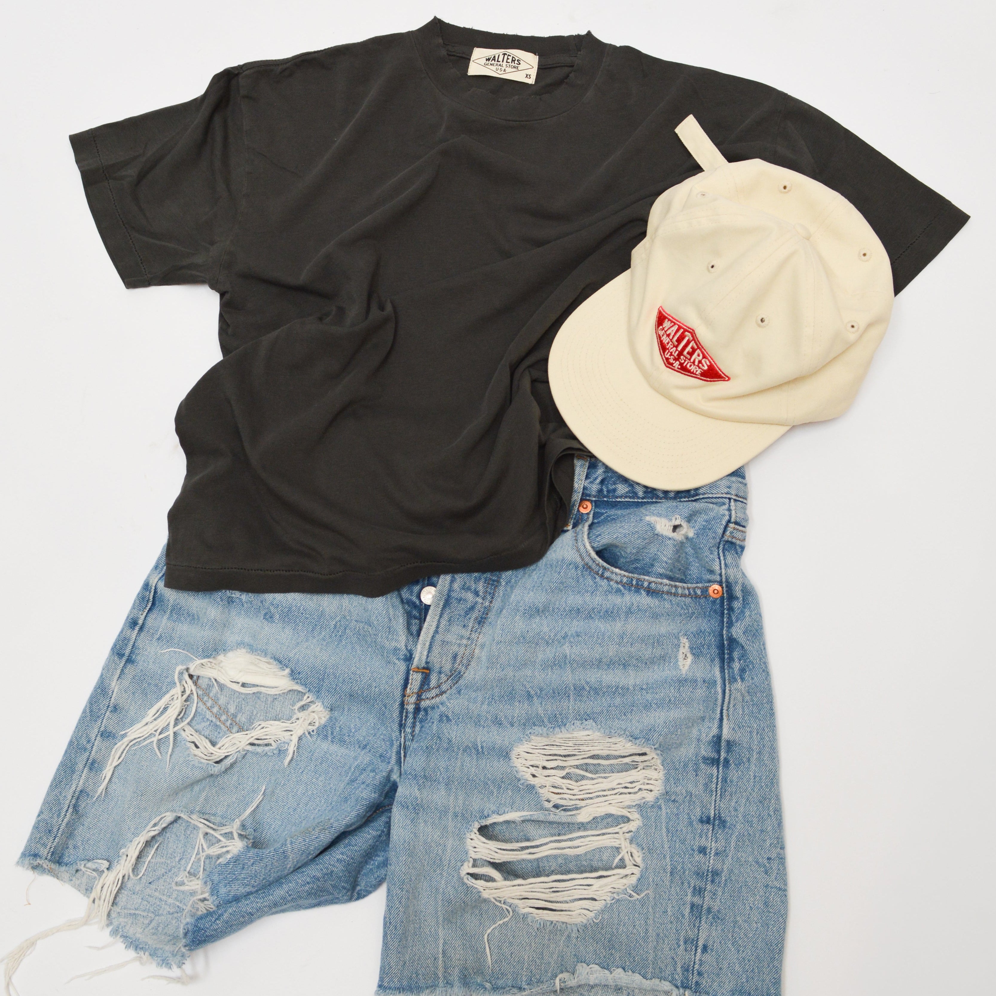 A styled flat-lay outfit featuring a black short-sleeve t-shirt, distressed light-wash blue denim shorts with ripped details, and a cream-colored baseball cap with a red logo, arranged on a clean white background.