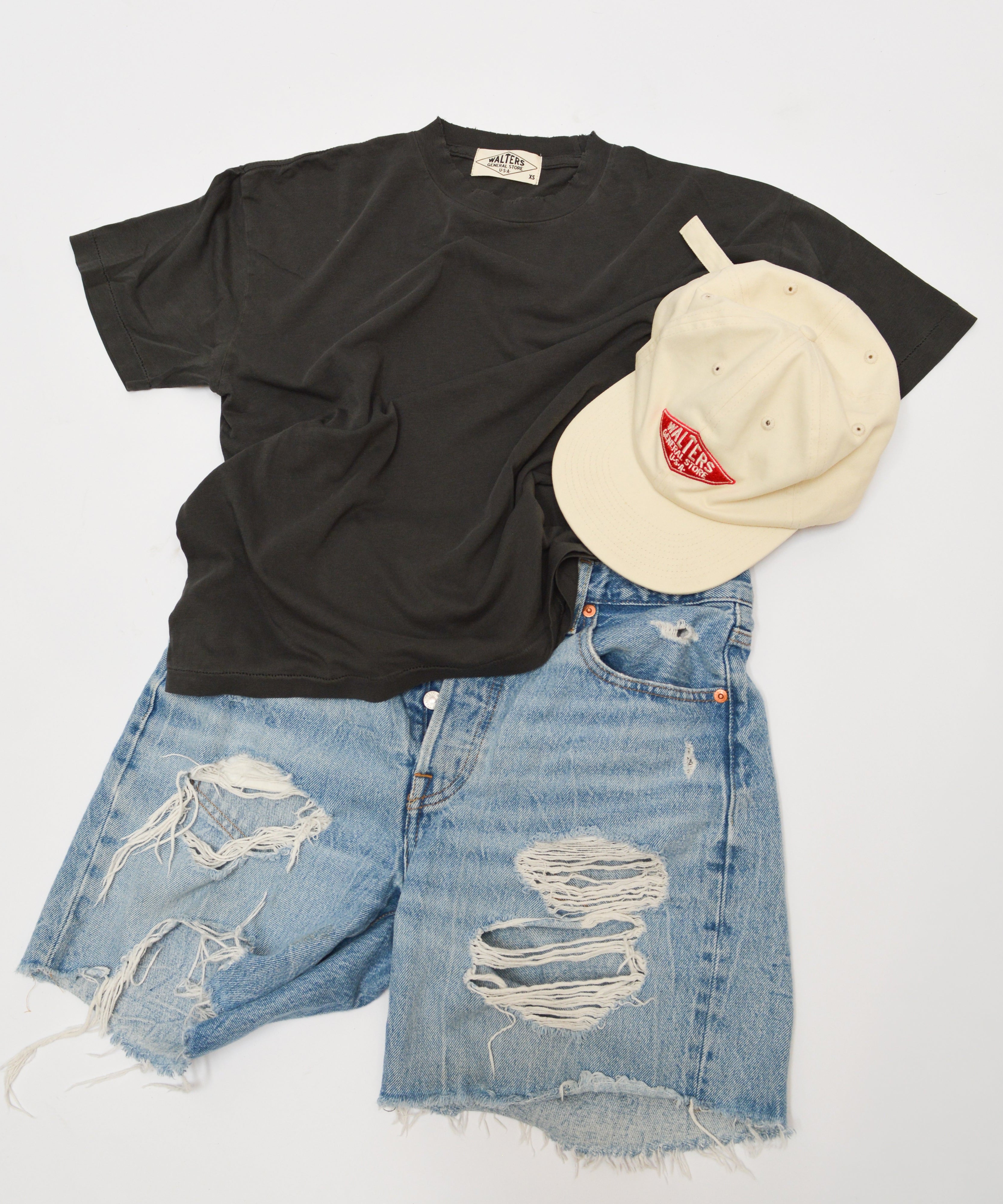 A styled flat-lay outfit featuring a black short-sleeve t-shirt, distressed light-wash blue denim shorts with ripped details, and a cream-colored baseball cap with a red logo, arranged on a clean white background.