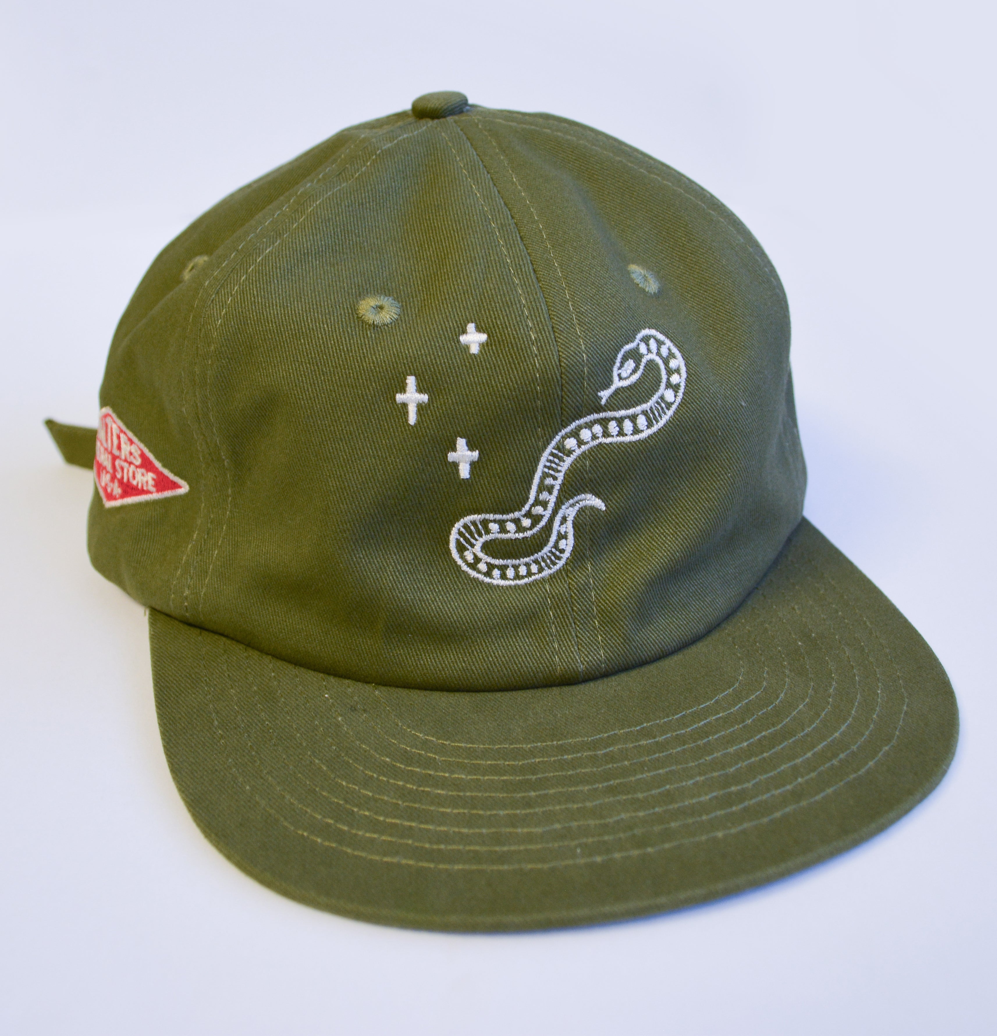 An olive green baseball cap featuring a white embroidered snake and plus-sign graphic on the front, with a red diamond patch on the side, set against a plain white background.