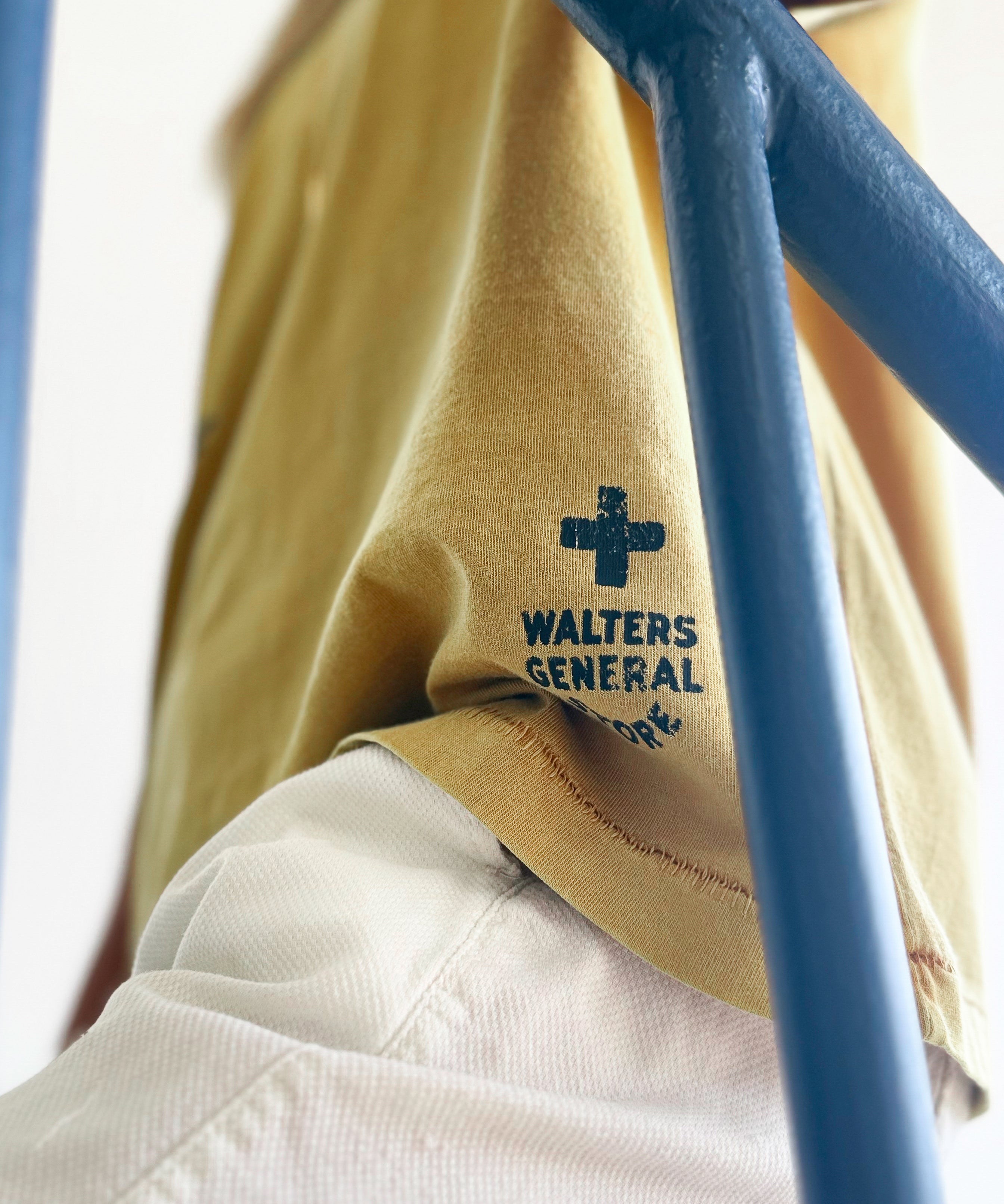A close-up lifestyle shot of a person wearing a mustard yellow t-shirt with a dark blue 'WALTERS GENERAL STORE' cross logo printed on the bottom left hem, paired with off-white trousers. A blue metal structure is in the foreground framing the shot.