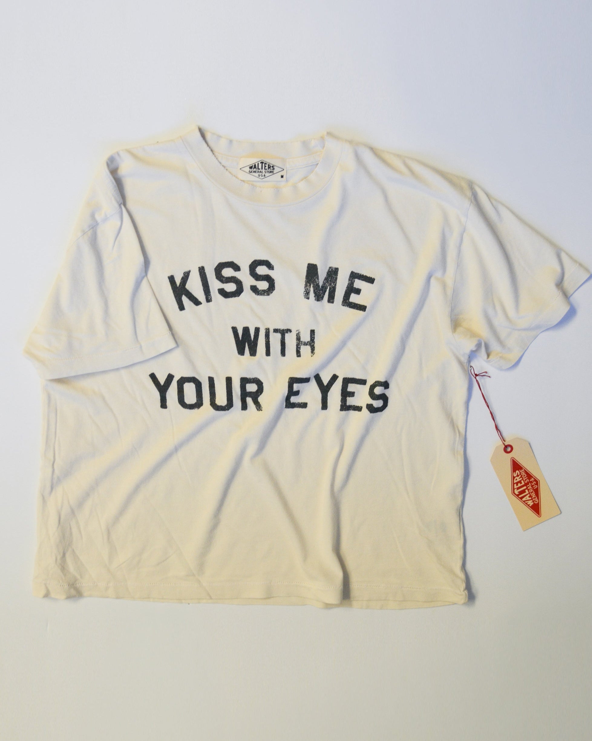 A cream-colored short-sleeve t-shirt with the text 'KISS ME WITH YOUR EYES' printed in black across the front. A small red and white Vans tag is attached to the right sleeve, and the shirt is laid flat against a light background.
