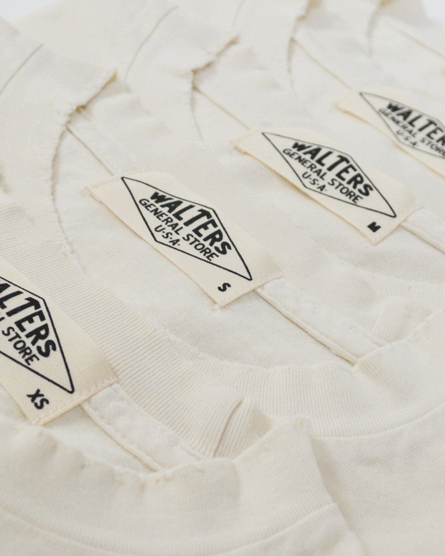 Three folded off-white cotton t-shirts stacked diagonally, showing the collar and printed 'Walters' brand labels.