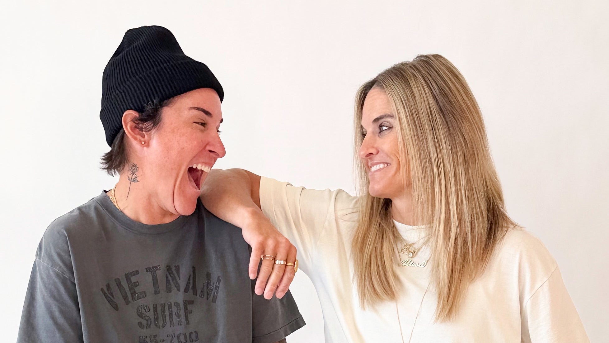 Two women laughing together against a plain white background. The woman on the left wears a black beanie, a gray graphic t-shirt, and olive green pants, while the woman on the right wears a cream-colored t-shirt and gold necklaces.