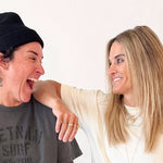 Two women laughing together against a plain white background. The woman on the left wears a black beanie, a gray graphic t-shirt, and olive green pants, while the woman on the right wears a cream-colored t-shirt and gold necklaces.