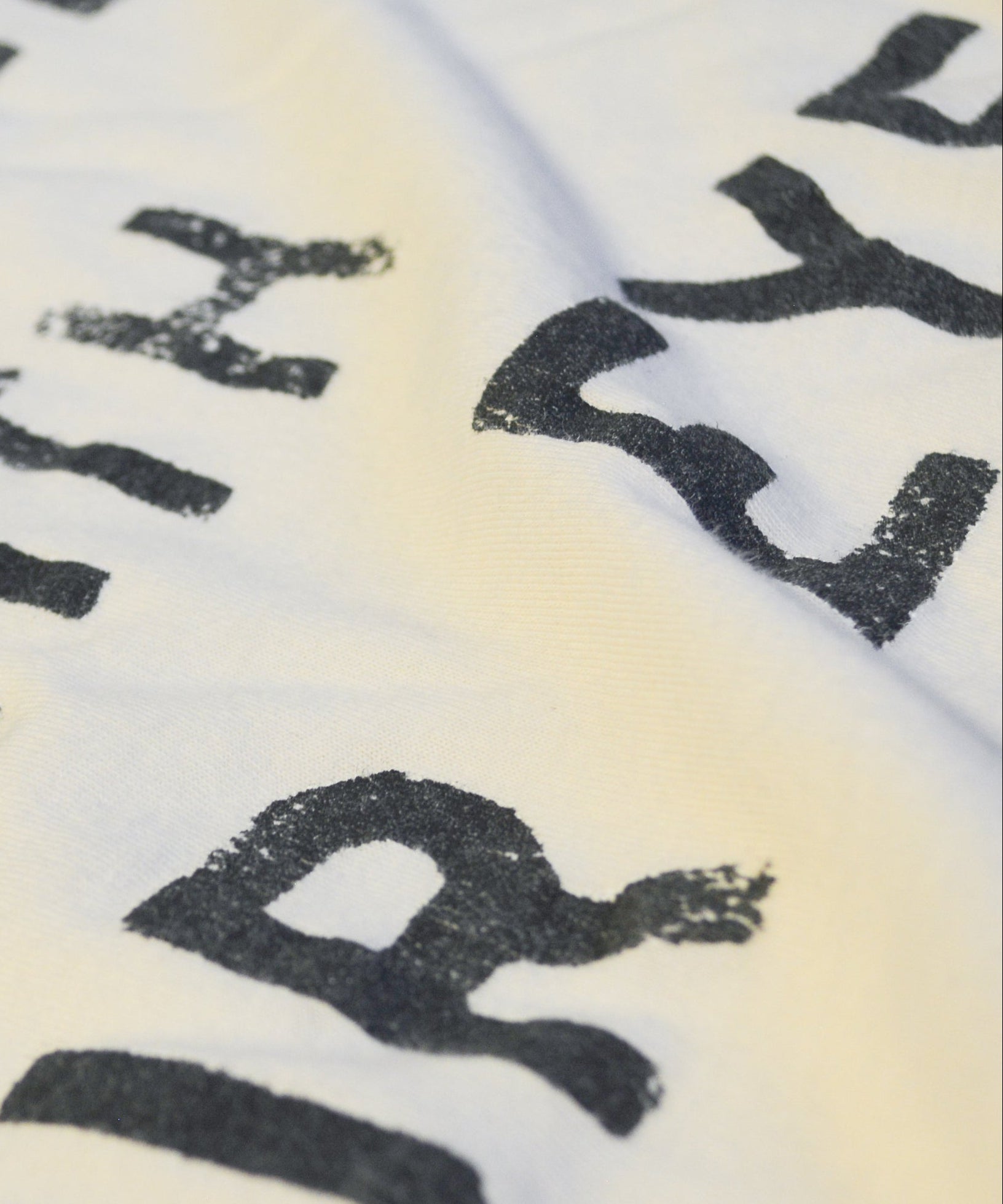 A close-up of a white fabric surface with large black distressed lettering partially visible, showing the texture and print detail.