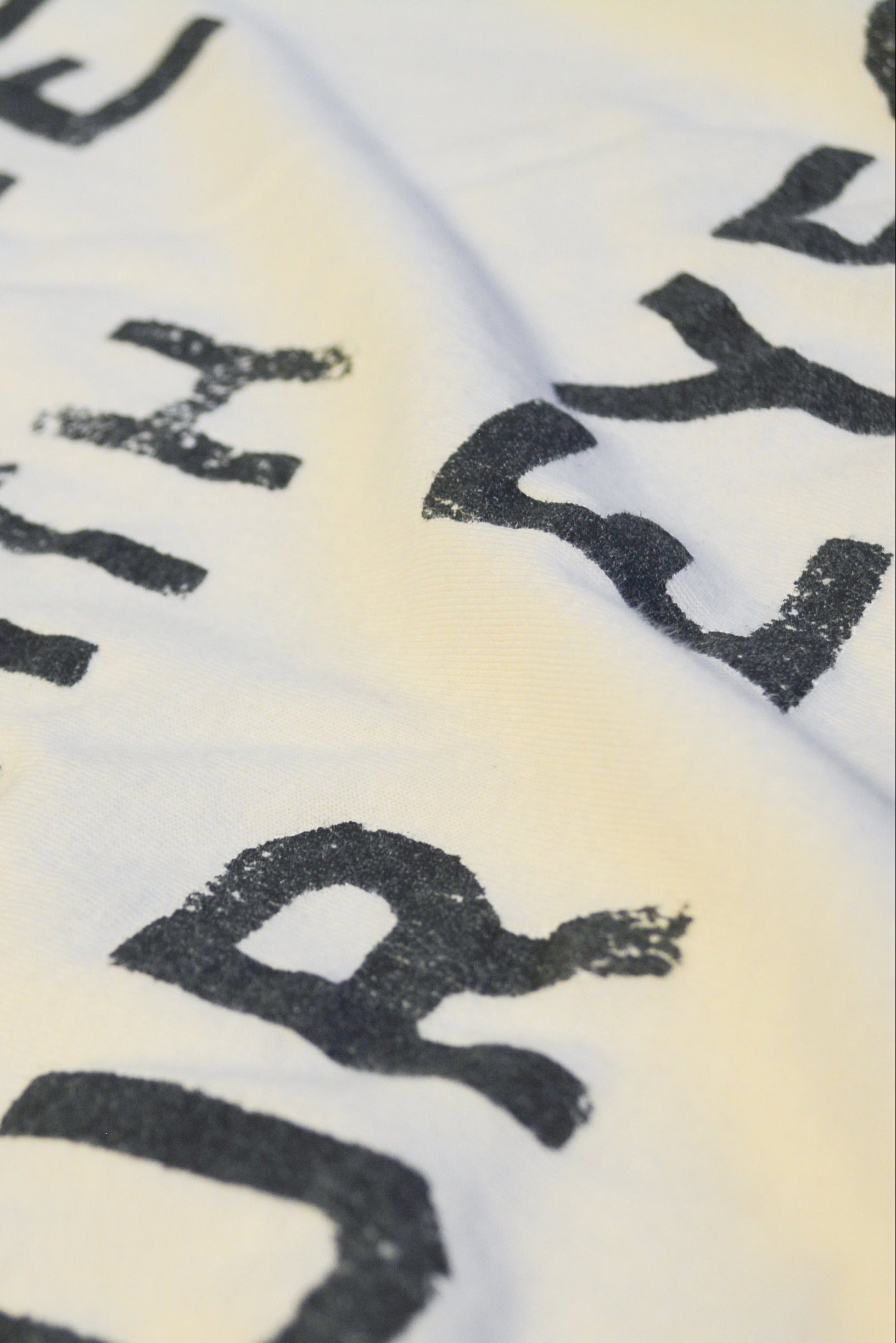 A close-up of a white fabric surface with large black distressed lettering partially visible, showing the texture and print detail.
