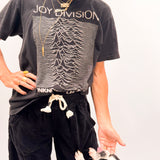 A woman wearing a black Joy Division graphic t-shirt, black sweatpants, and a khaki hat is standing next to a black and white dog on a white studio background.