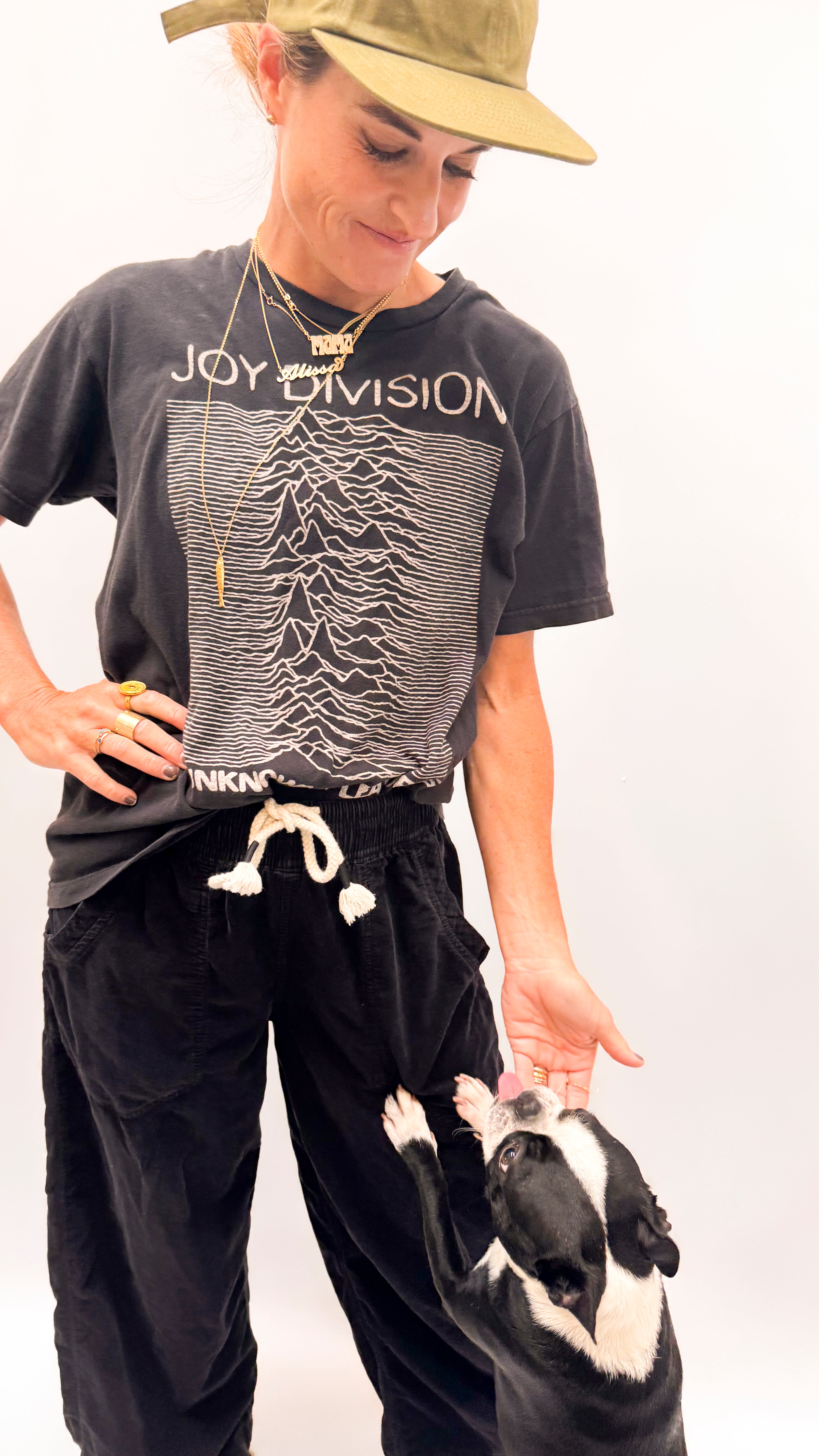 A woman wearing a black Joy Division graphic t-shirt, black sweatpants, and a khaki hat is standing next to a black and white dog on a white studio background.