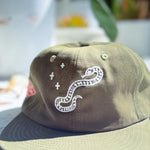 An olive green cap with white embroidered details, featuring a snake and plus-sign motif on the front panel, photographed in a bright, slightly overexposed outdoor setting.