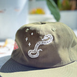 An olive green cap with white embroidered details, featuring a snake and plus-sign motif on the front panel, photographed in a bright, slightly overexposed outdoor setting.