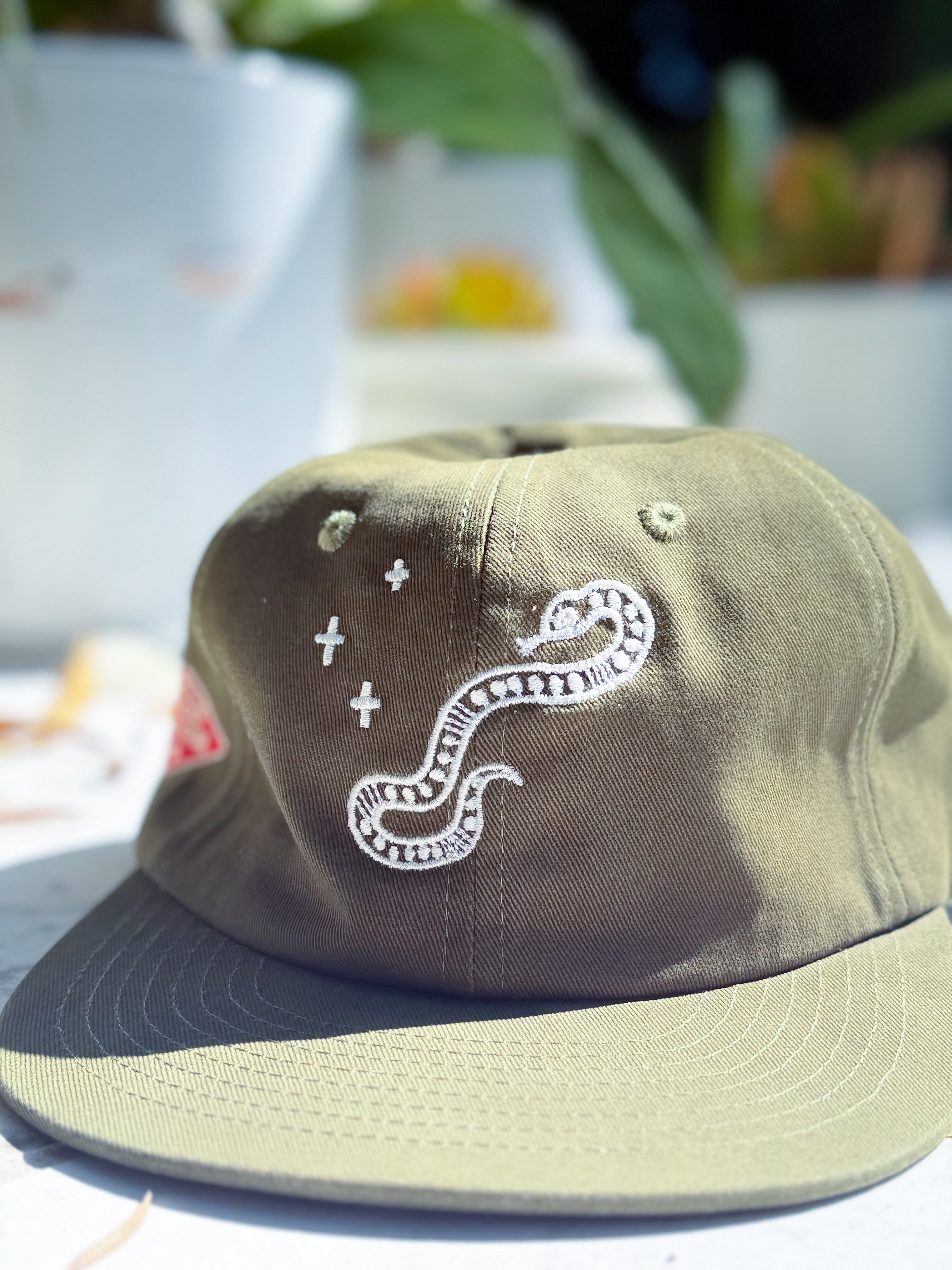 An olive green cap with white embroidered details, featuring a snake and plus-sign motif on the front panel, photographed in a bright, slightly overexposed outdoor setting.