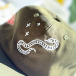 Close-up of an olive green baseball cap featuring white embroidered details, including a snake motif and small cross symbols.