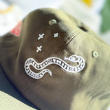 Close-up of an olive green baseball cap featuring white embroidered details, including a snake motif and small cross symbols.