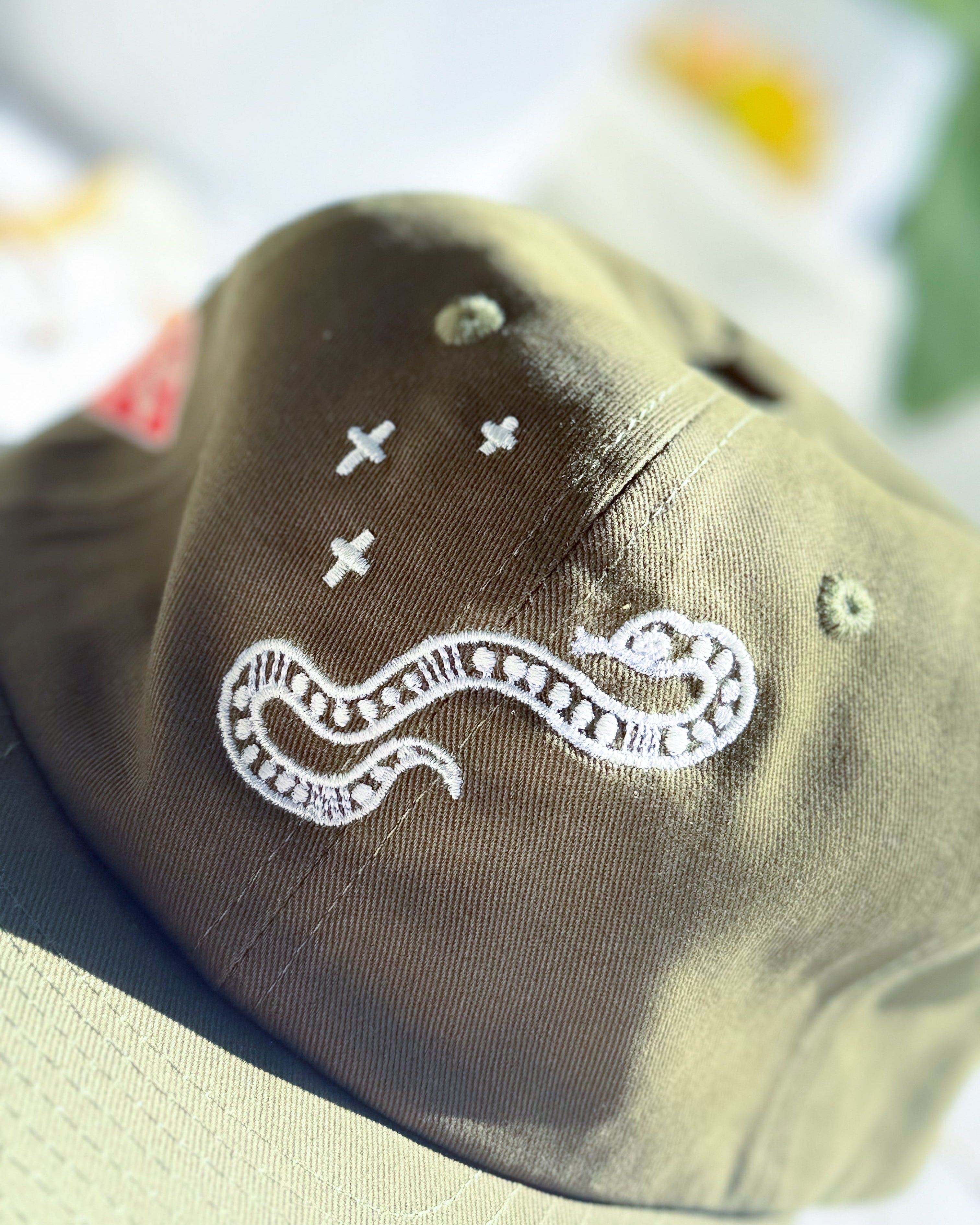 Close-up of an olive green baseball cap featuring white embroidered details, including a snake motif and small cross symbols.