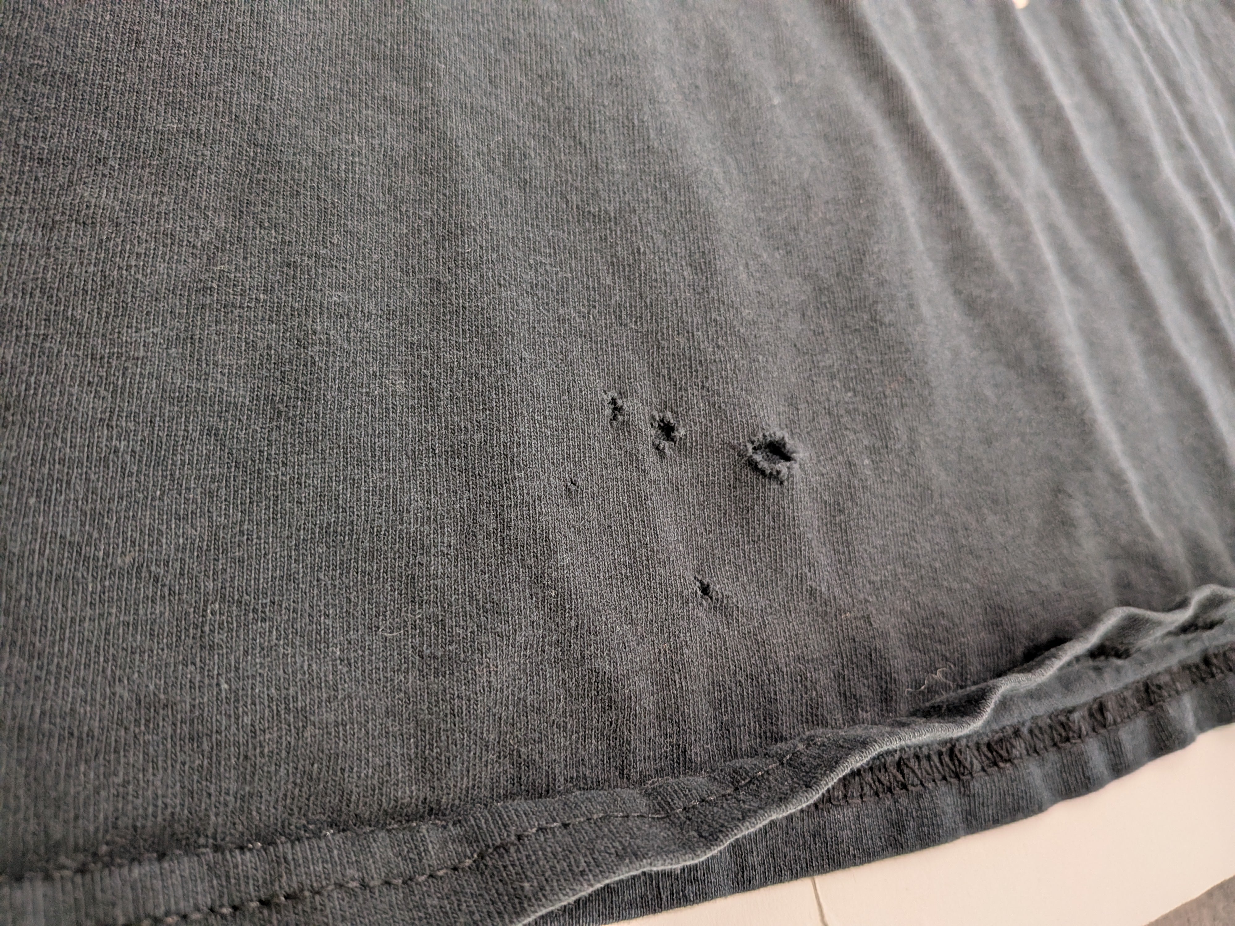 Close-up of a dark gray washed cotton fabric, showing a hemmed edge with visible frayed threads and small distressed holes in the knit texture.