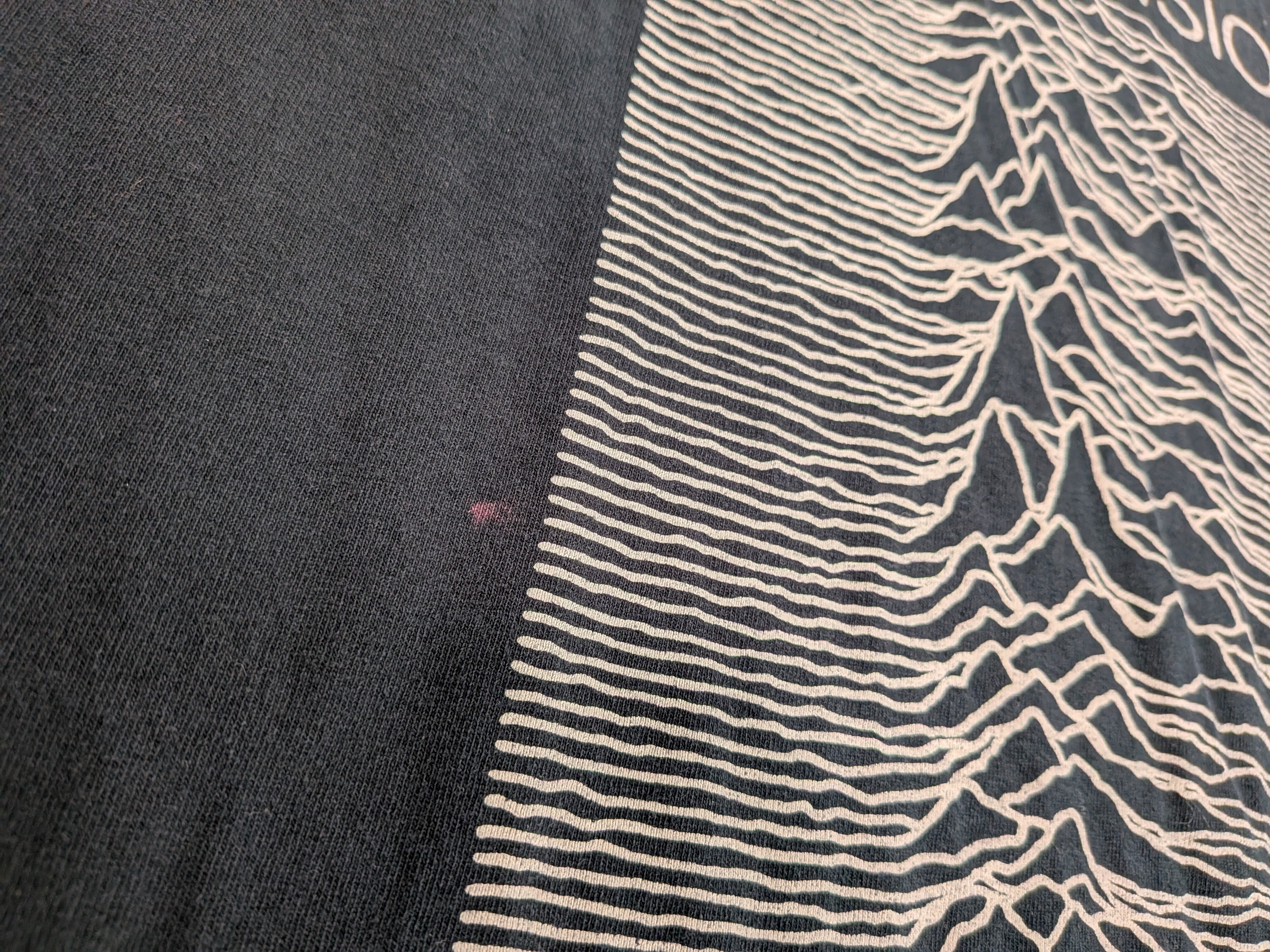 Close-up of a black knit garment featuring a cream-colored graphic print of the album 'Unknown Pleasures' wave pattern.