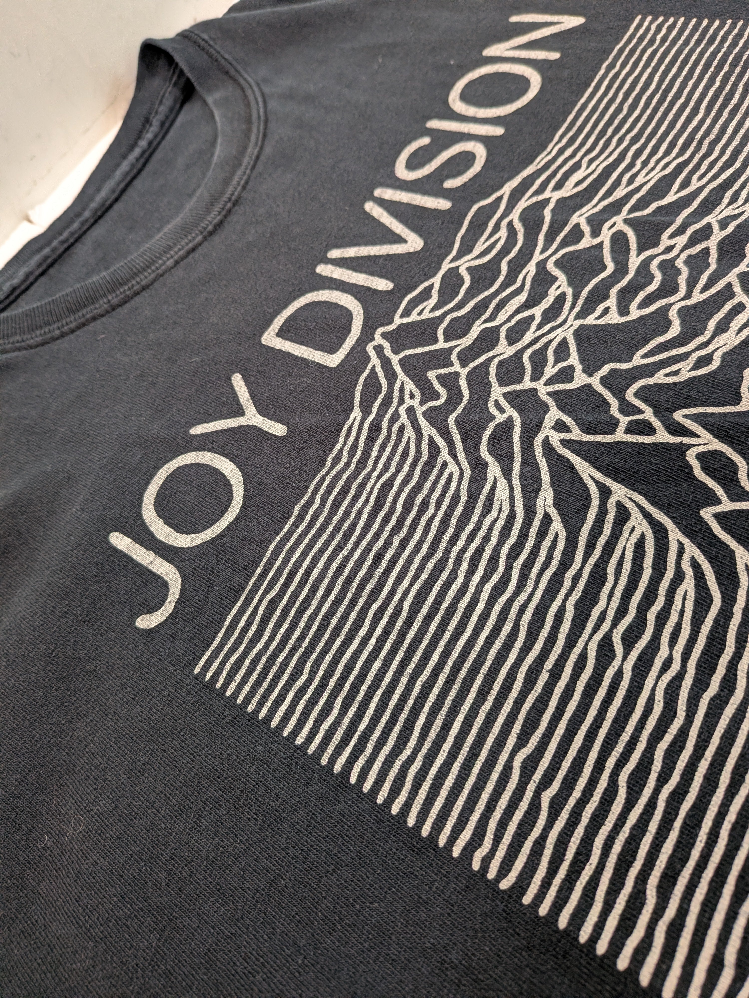 Close-up of a black Joy Division graphic t-shirt featuring the band's name and the 'Unknown Pleasures' line-art print.